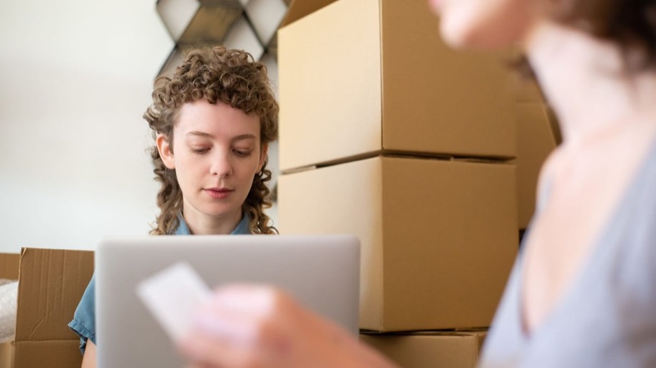 Two people working at a laptop among stacked shipping boxes